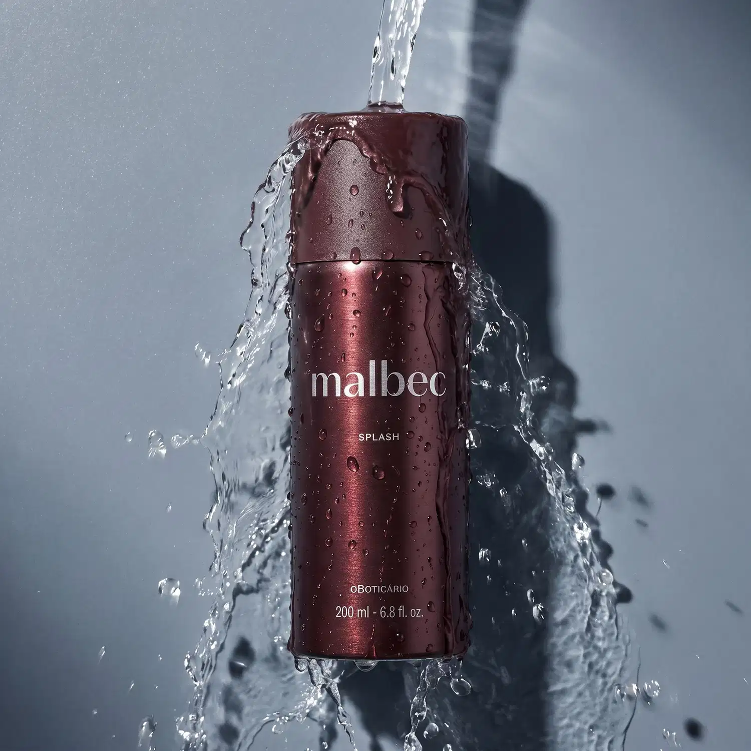 Frasco marrom cilíndrico de item de perfumaria da marca Malbec sob jato d'água, com gotas de água sobre a superfície cinza clara.