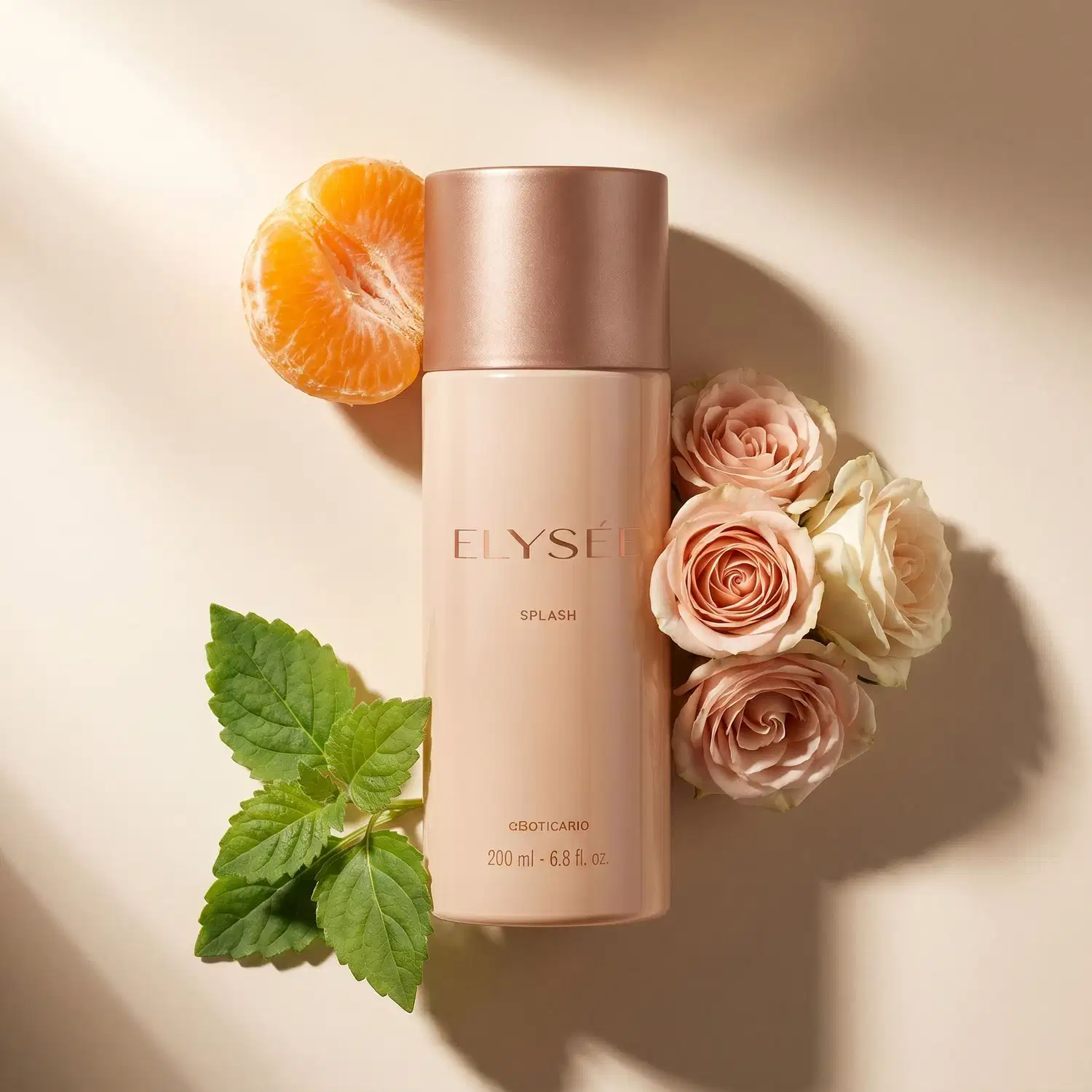 Frasco de body splash da marca Elysée na cor rosa claro com tampa na cor rosé gold, cercado por rosas, meia tangerina descascada e folhas de menta sobre superfície rosa clara e iluminação clara.