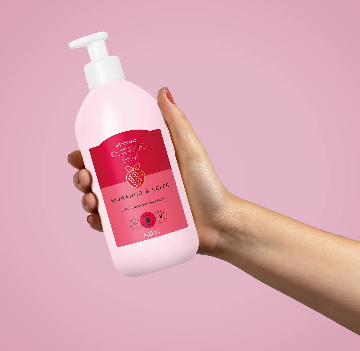 Imagem de uma embalagem branca de loção hidratante corporal Cuide-se Bem Morango e Leite com rótulo vermelho e rosa e imagem de morango, segura por mão feminina contra fundo rosa.