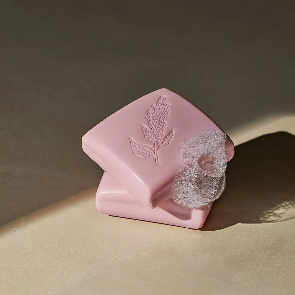 Imagem de dois sabonetes quadrados rosa, um em cima do outro, em superfície bege com sombreado, contendo espuma branca em cima deles. O ambiente sugere um contexto de relaxamento e cuidado pessoais.