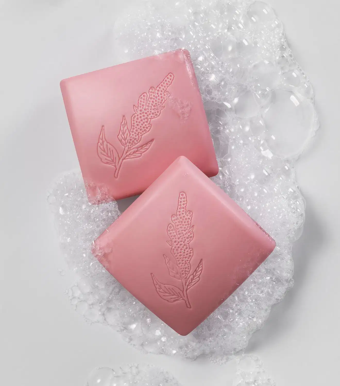 Imagem de dois sabonetes quadrados na cor rosa da marca Nativa SPA Ameixa Plum em superfície molhada com espuma branca ao redor dos sabonetes.