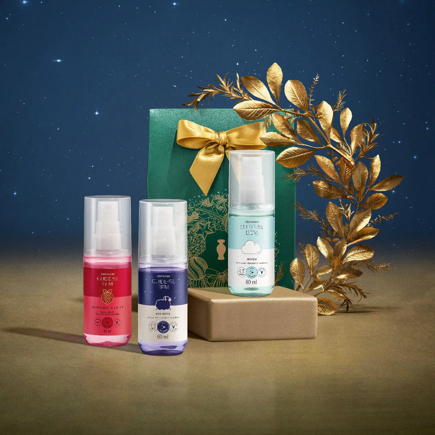 Três frascos de itens de mini body splashes da marca Cuide-se Bem e uma caixa de presente verde com laço dourado em cenário de céu noturno estrelado sobre superfície bege e um adorno de folhas douradas ao redor da caixa de presente.
