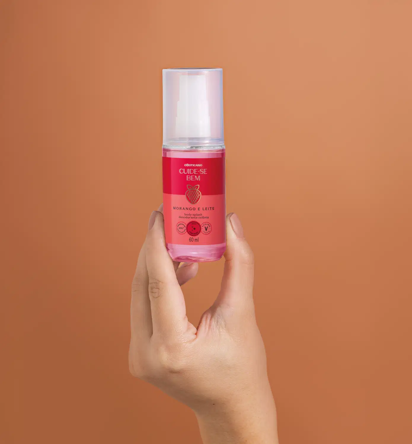 Imagem de uma mão feminina segurando um vidro transparente com líquido rosa, borrifador branco e tampa transparente contendo body splash Cuide-se Bem Morango e Leite. O fundo é rosa vibrante.