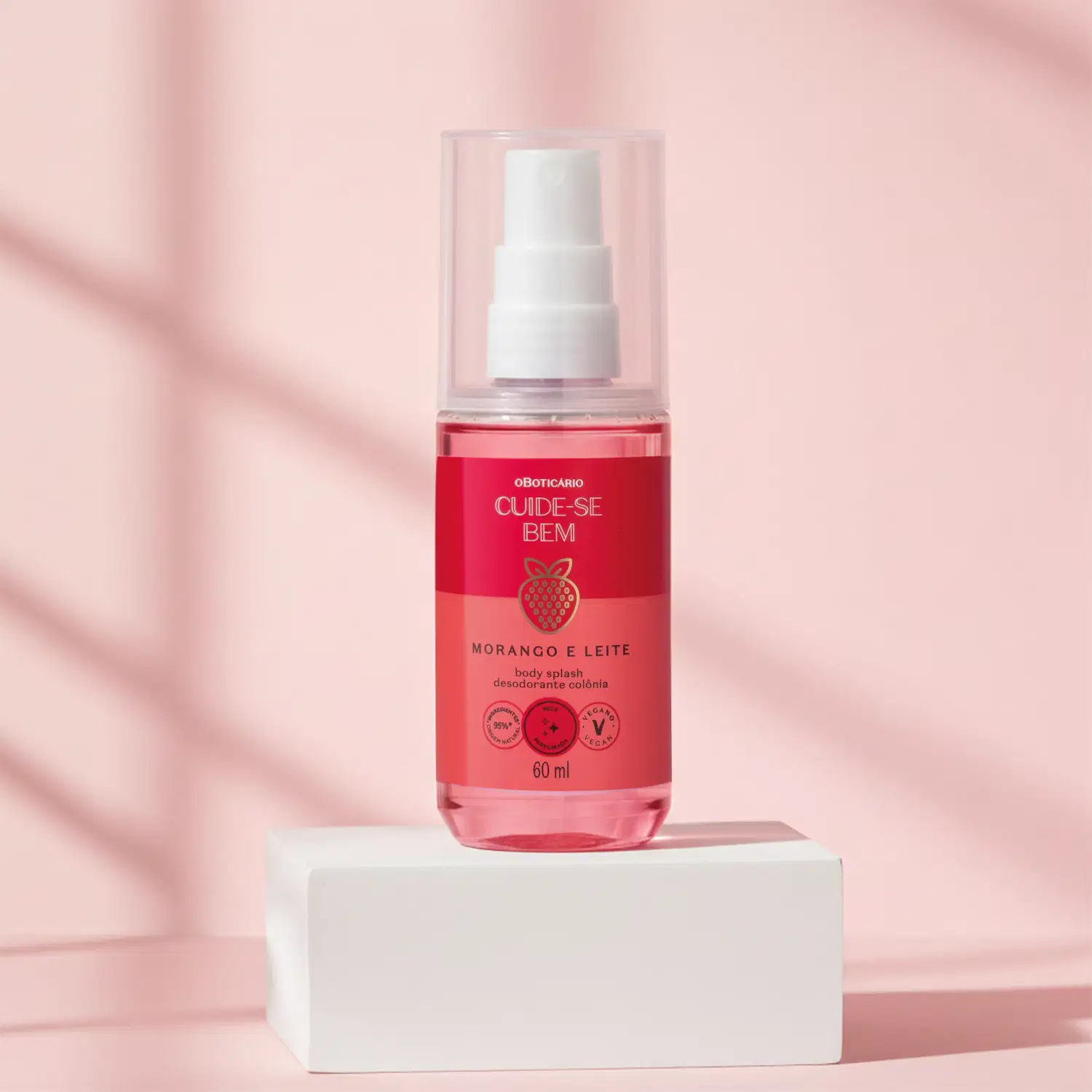 Imagem de um frasco transparente com rótulo vermelho e rosa, líquido rosa, borrifador branco e tampa transparente de body splash Cuide-se Bem Morango e Leite em cima de uma superfície branca, com fundo rosa-claro e sombras suaves.