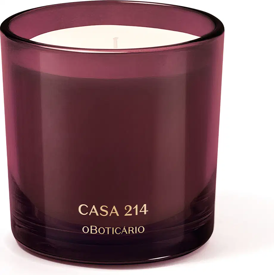 Vela aromática em recipiente de vidro de cor vinho com rótulo "Casa 214 o Boticario" em letras douradas. O recipiente possui formato cilíndrico e a vela em seu interior parece ser de cor clara.