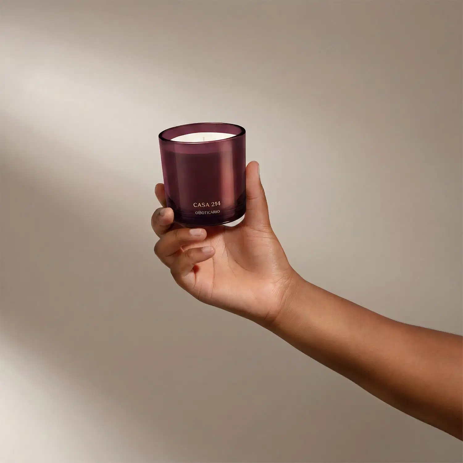 Vela aromática em recipiente de vidro de cor vinho com rótulo "Casa 214 o Boticario" em letras douradas. O recipiente possui formato cilíndrico e a vela em seu interior parece ser de cor clara.