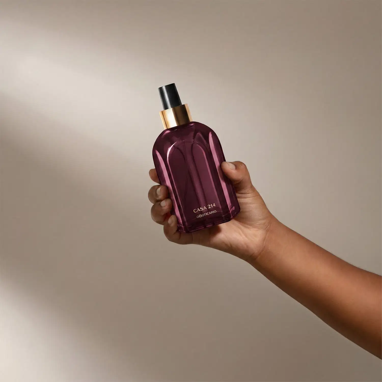 Braço com mão segurando um vidro roxo do aromatizador de ambientes Casa 214 Figo Magnífico. Ele possui válvula spray preta e rótulo em dourado. O fundo é neutro, criando um ambiente minimalista e elegante.