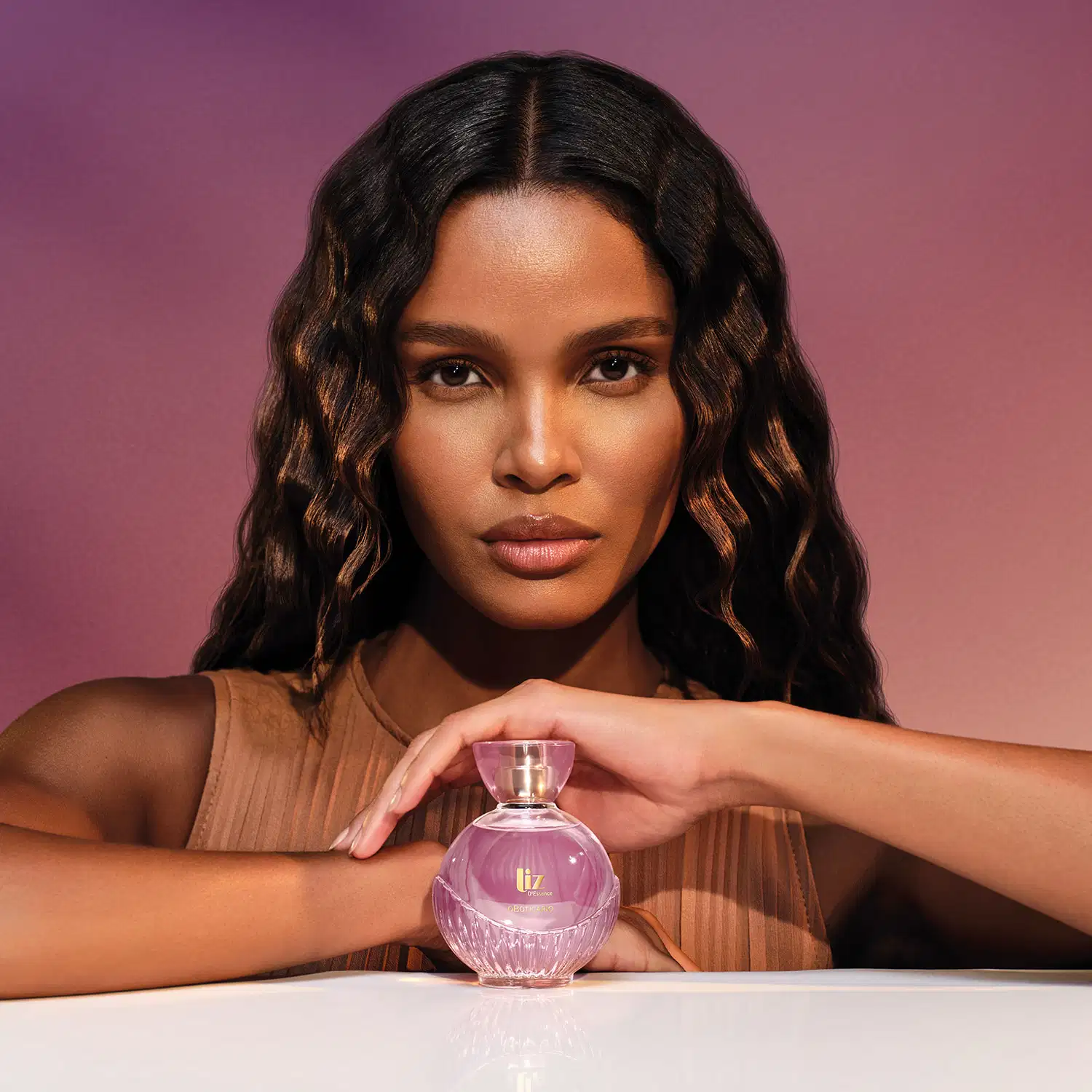 Modelo negra com cabelos longos e ondulados e expressão séria vista dos ombros para cima. A frente dela, um frasco de desodorante colônia da marca Liz D'Essence, sobre o qual ela repousa sua mão esquerda.