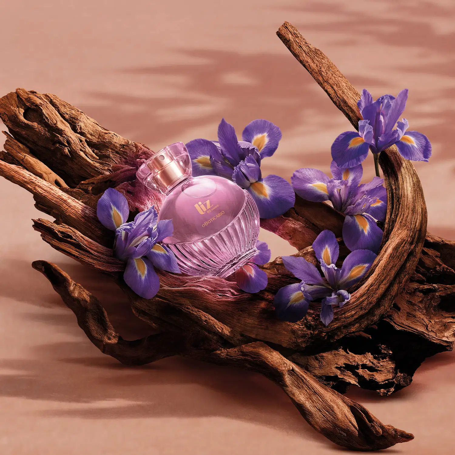 Ambiente rústico e natural com flores de íris violetas, galhos de madeira e um frasco de perfume da marca Liz D'Essence, em tons de rosa, criando uma atmosfera elegante e delicada.