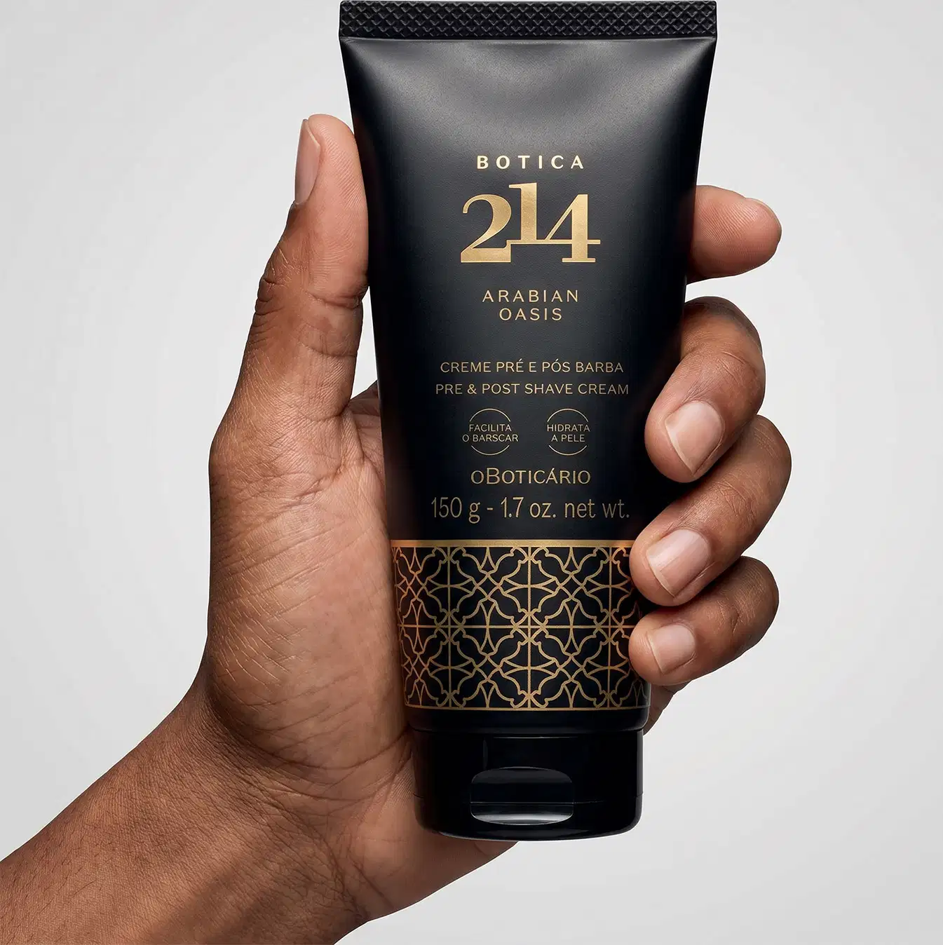 Tubo preto com texto e arabescos dourados da marca Botica 214 Arabian Oasis em fundo branco, sendo segurado por mão masculina de pele parda.