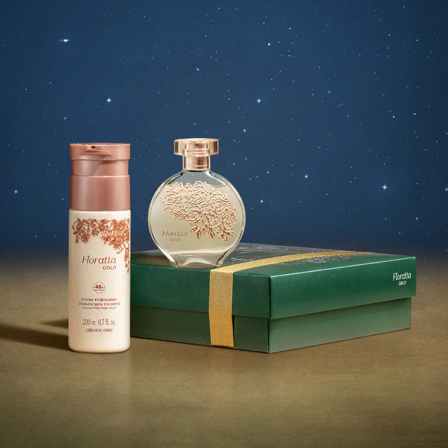 Frasco de vidro de desodorante colônia Floratta Gold sobre  caixa presenteável verde com ilustração natalina. Ao lado um frasco branco com tampa dourada de body splash da mesma linha sobre superfície marrom e fundo azul-escuro simulando um céu estrelado.