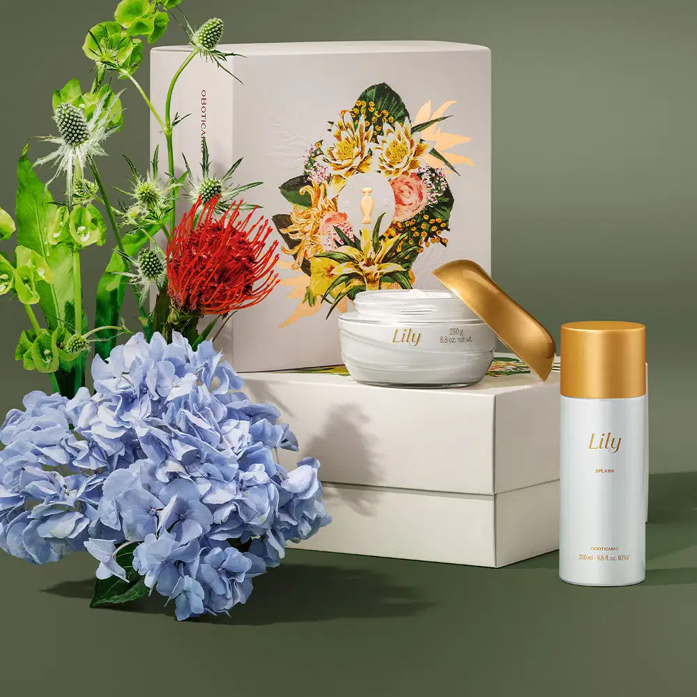Imagem do kit de cuidados Lily, com pote de creme acetinado corporal e um frasco de body splash, além de caixa com ilustração floral. Ao lado, flores azuis, vermelhas e brancas, sobre superfície cinza.