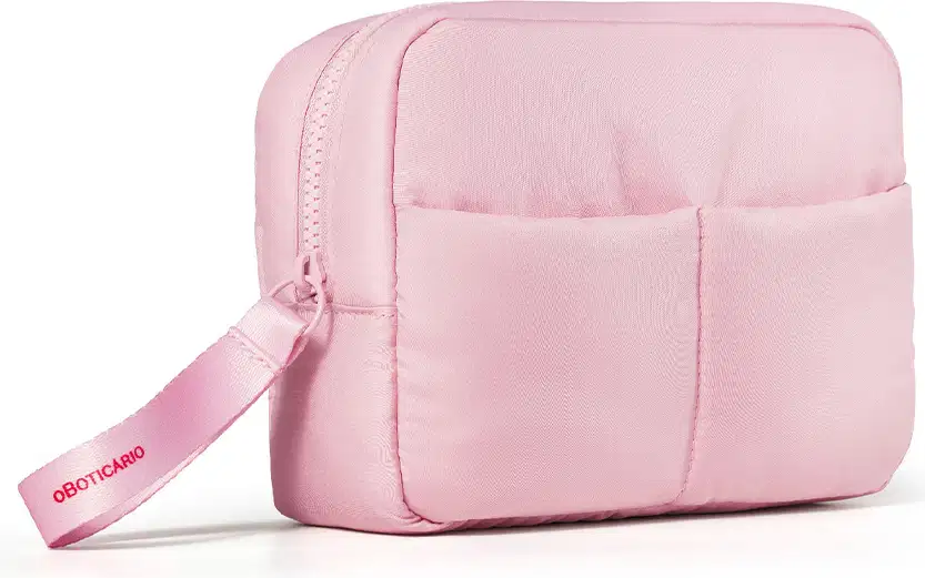 Imagem de uma nécessaire rosa clara com puxador. A bolsa tem vários compartimentos e um zíper na parte frontal. Há o nome da marca o Boticário em rosa escuro no puxador.