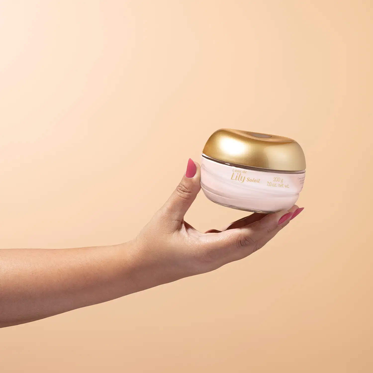 Imagem de uma mão feminina segurando um pote de vidro transparente com conteúdo branco e tampa dourada contendo creme acetinado corporal da marca L'eau de Lily Soleil em fundo bege vibrante.