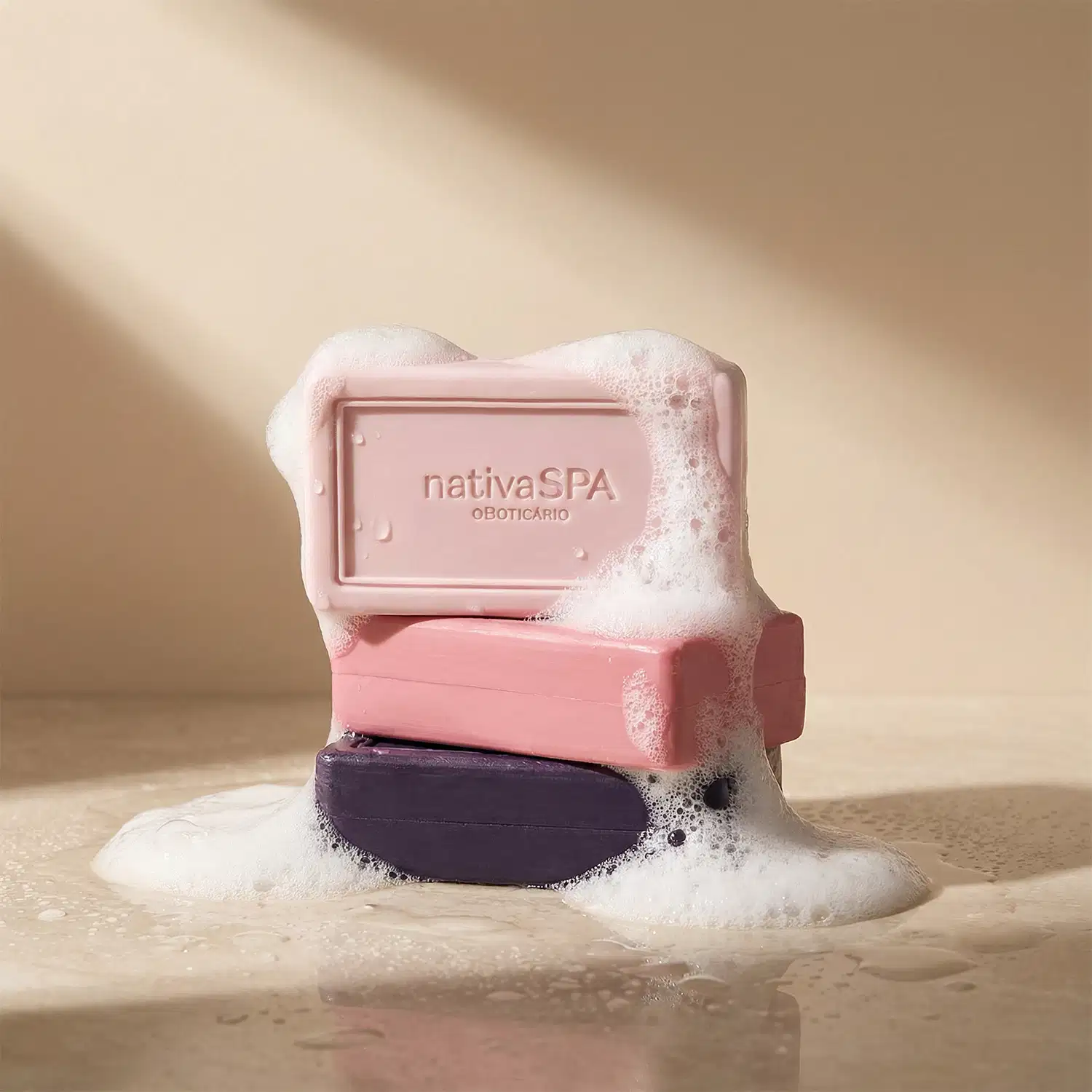 A imagem mostra três barras de sabonete em tons de rosa e roxo da marca Nativa SPA estão cheios de espuma branca, sobre superfície clara, sombreada e molhada.