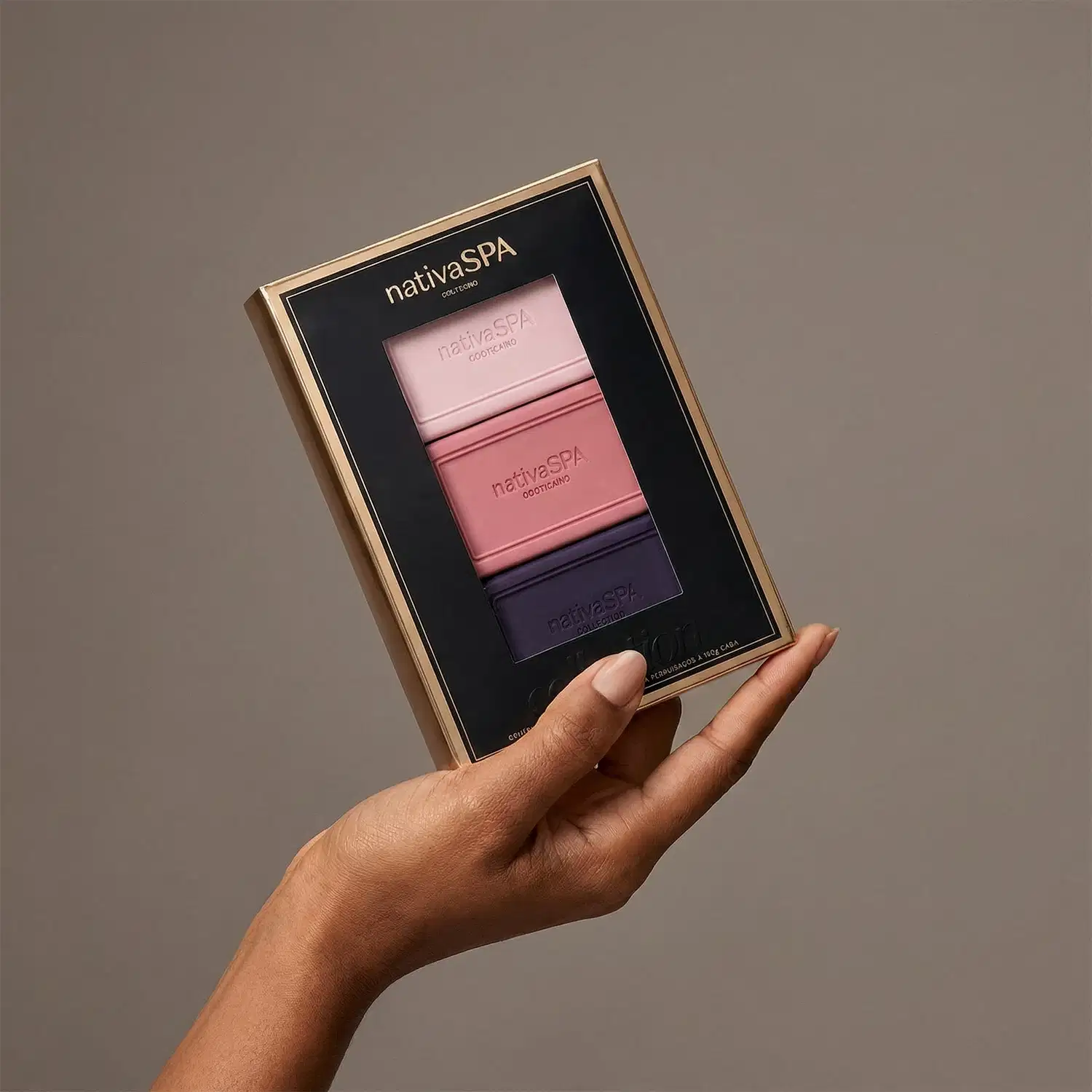 A imagem mostra um braço com mão segurando uma caixa de presente retangular preta com detalhes dourados, contendo três sabonetes em barra da marca Nativa SPA em tons de rosa e roxo. O fundo é neutro, sóbrio e minimalista.
