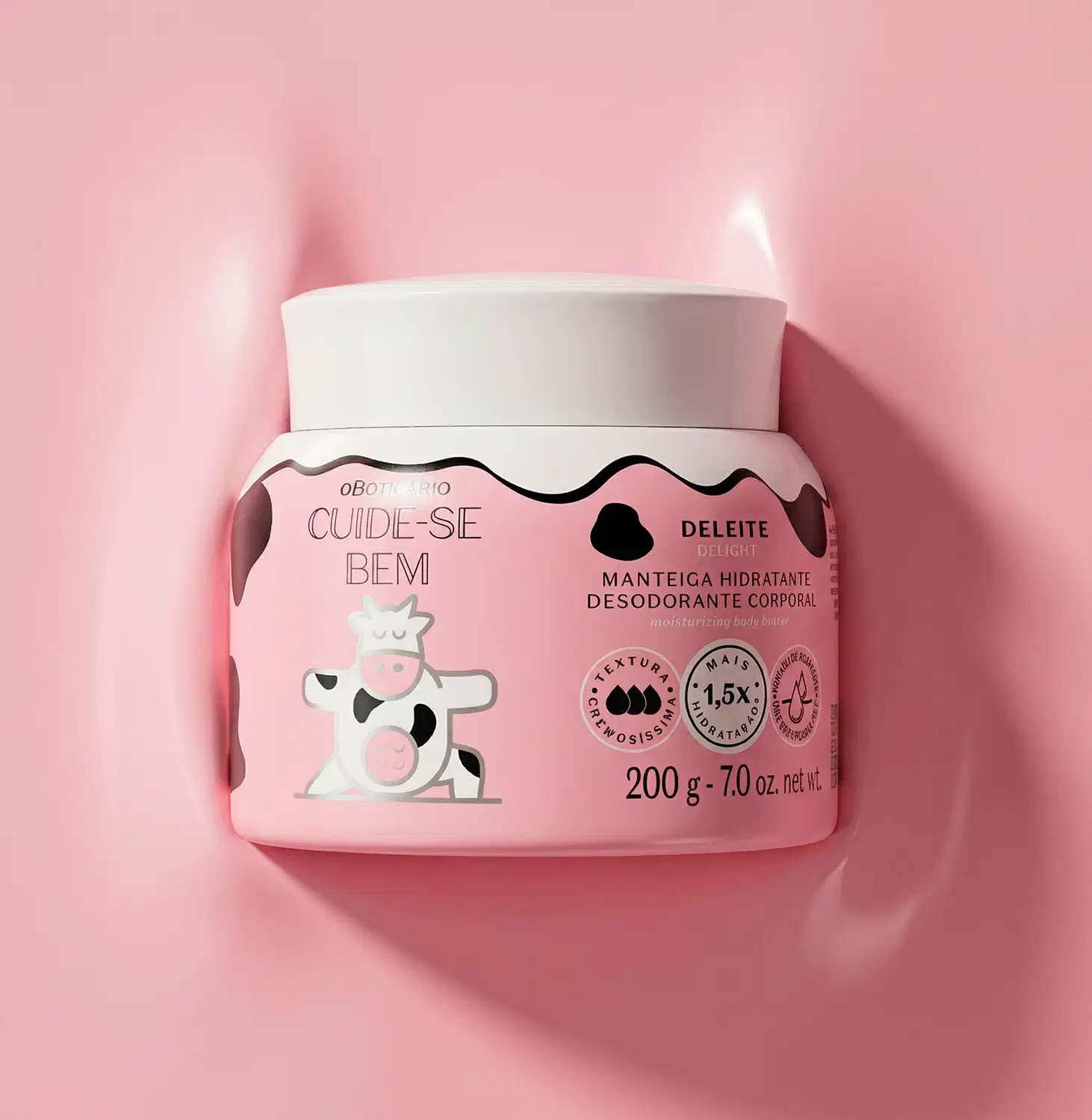 A imagem mostra o pote branco de manteiga corporal Cuide-se Bem Deleite deitado em uma textura cremosa na cor rosa. A embalagem é branca com rótulo em rosa e estampa de vaca. Ele tem tampa branca e contém 200g de produto.