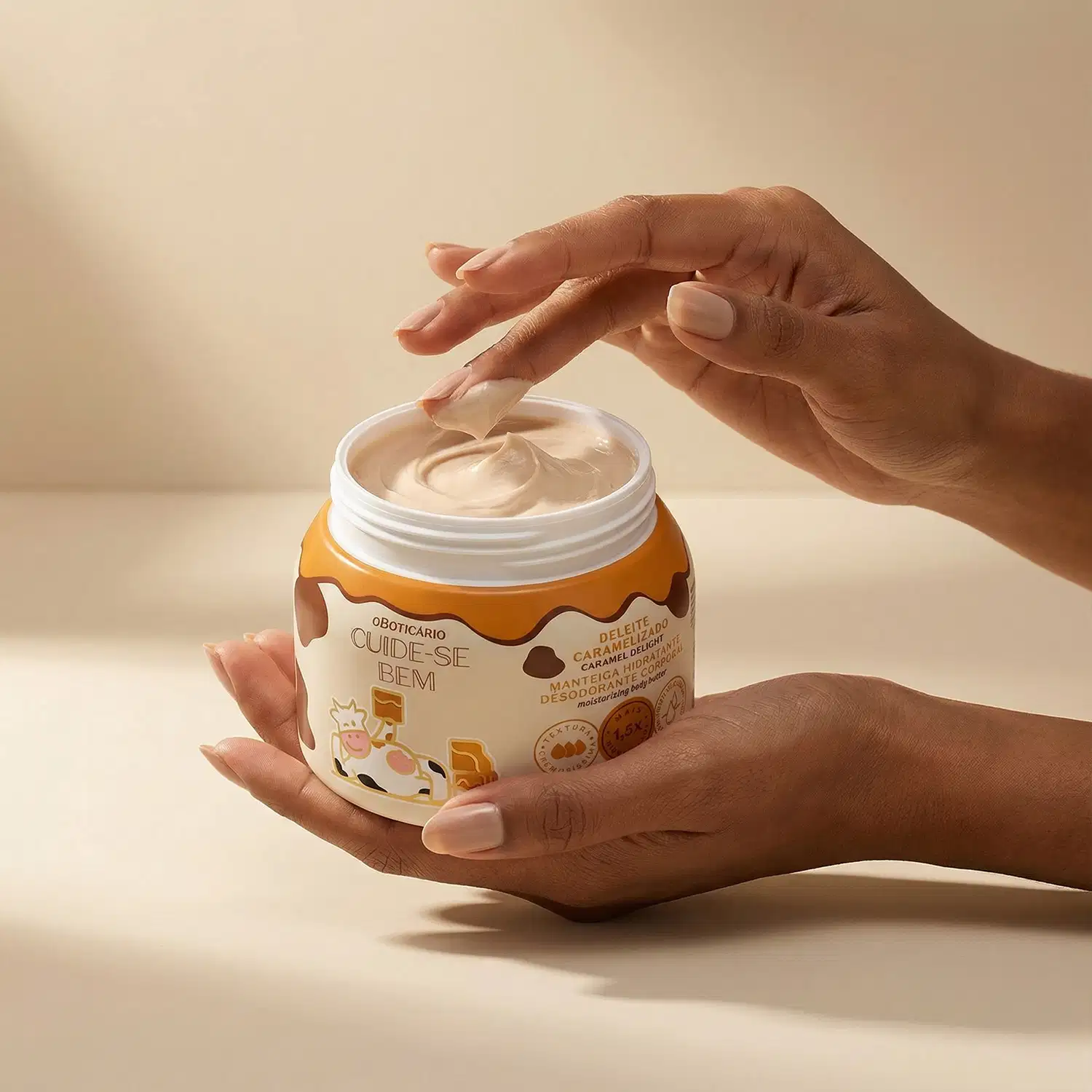 A imagem mostra o recipiente está sendo segurado por mãos femininas e os dedos estão cheios de manteiga corporal, apresentando a textura do produto. O ambiente é neutro, com fundo bege claro, sugerindo um contexto de cuidados pessoais.
