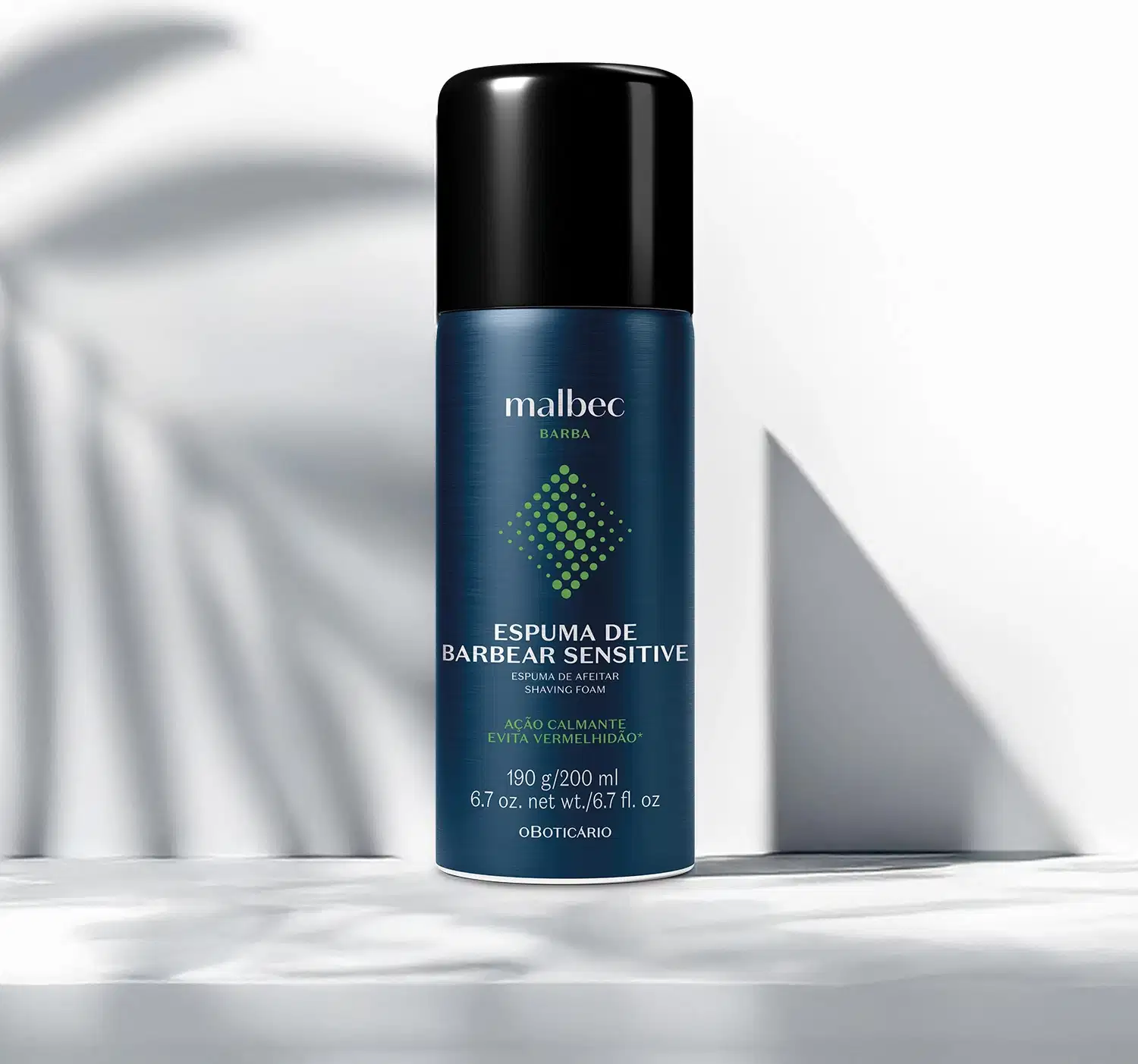 A embalagem verde metálica da espuma de barbear da marca Malbec está sobre superfície clara com sombras suaves, em ambiente minimalista. O produto possui rótulo em branco e dourado e tampa preta.