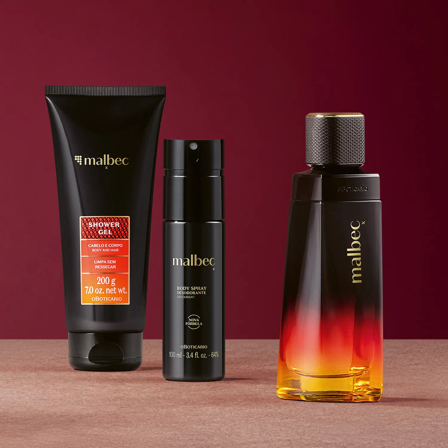 Três produtos de perfumaria da linha Malbec X em uma superfície rosa e fundo vinho, incluindo um frasco de item de perfumaria, um body spray e um gel de banho em embalagem preta e laranja.