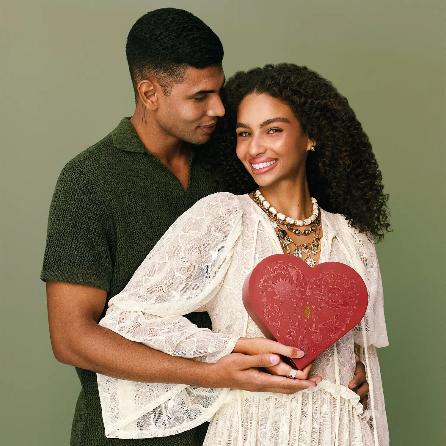 Casal negro jovem em fundo cinza, sendo que a mulher veste vestido branco de manga longa esvoaçante e segura uma caixa de presente em formato de coração vermelho enquanto o homem veste camiseta verde escuro e a abraça carinhosamente.