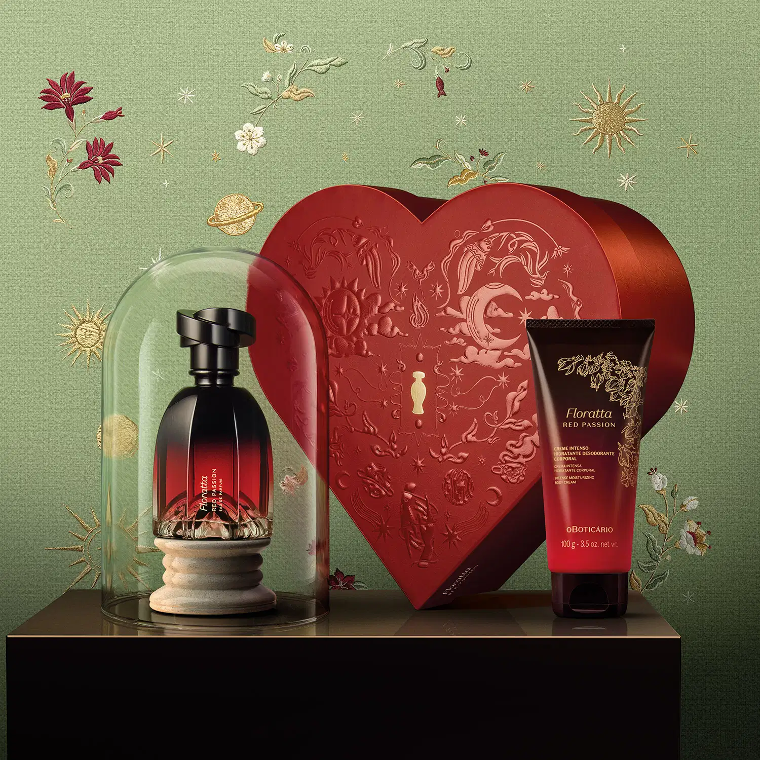 Cena de fundo verde com flores e estrelas douradas bordadas ao fundo. No primeiro plano, uma superfície marrom exibindo um frasco Eau de Parfum Floratta Red Passion dentro de uma redoma de vidro, uma bisnaga de creme corporal e caixa em formato de coração