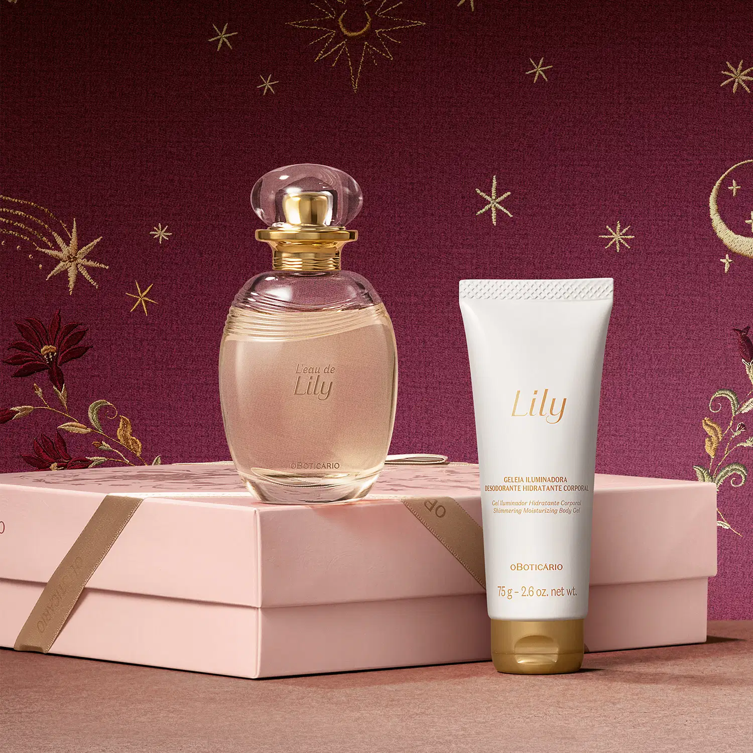 Embalagem rosa posicionada horizontalmente sobre superfície rosa com frasco de item de perfumaria da marca L'Eau de Lily sobre ela e bisnaga de creme corporal branca ao lado, sobre fundo bordô com estrelas douradas.