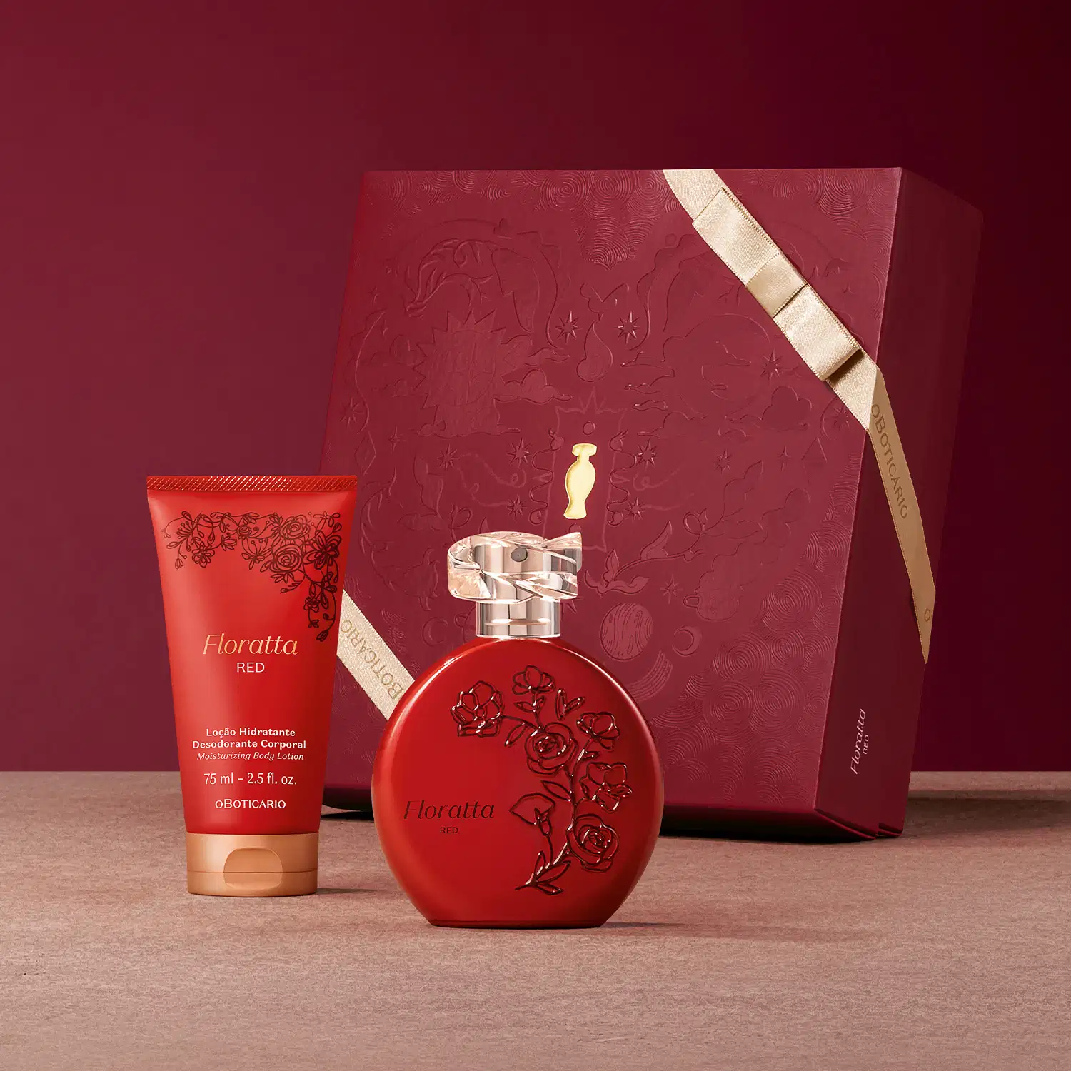 Em primeiro plano, frasco vermelho com detalhes florais em relevo e bisnaga de loção hidratante desodorante, ambos da linha Floratta Red sobre superficie rosa clara. Ao fundo caixa de presente vermelha com textura e laço dourado em fundo vinho.