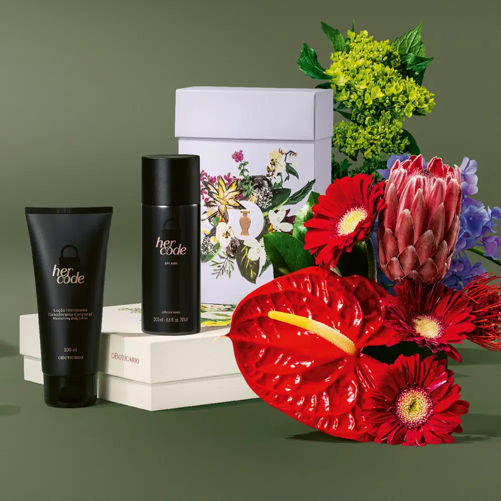 Imagem de produtos da marca Her Code, sendo um frasco de body splash e um tubo de Loção Hidratante Corporal, além de uma caixa decorada com flores. Ao lado do kit, flores vermelhas, amarelas e roxas em arranjo sobre superfície verde.