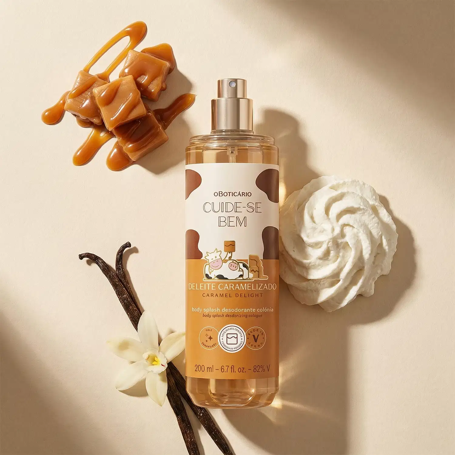 Recipiente de body splash Cuide-se Bem Deleite Caramelizado em formato de spray, de rótulo branco com detalhes em marrom e imagem de vaca. Ao redor, há pedaços de caramelo, uma flor branca e um creme branco, em um ambiente com tons neutros.