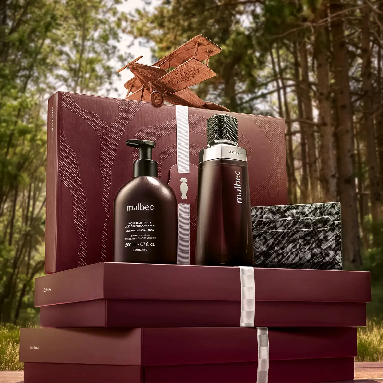Caixa de presente com produtos de beleza da marca Malbec, em ambiente externo com vegetação ao fundo. Embalagem em tons de vermelho e preto, com desodorante, loção hidratante e perfume, sugerindo um conjunto de cuidados pessoais.