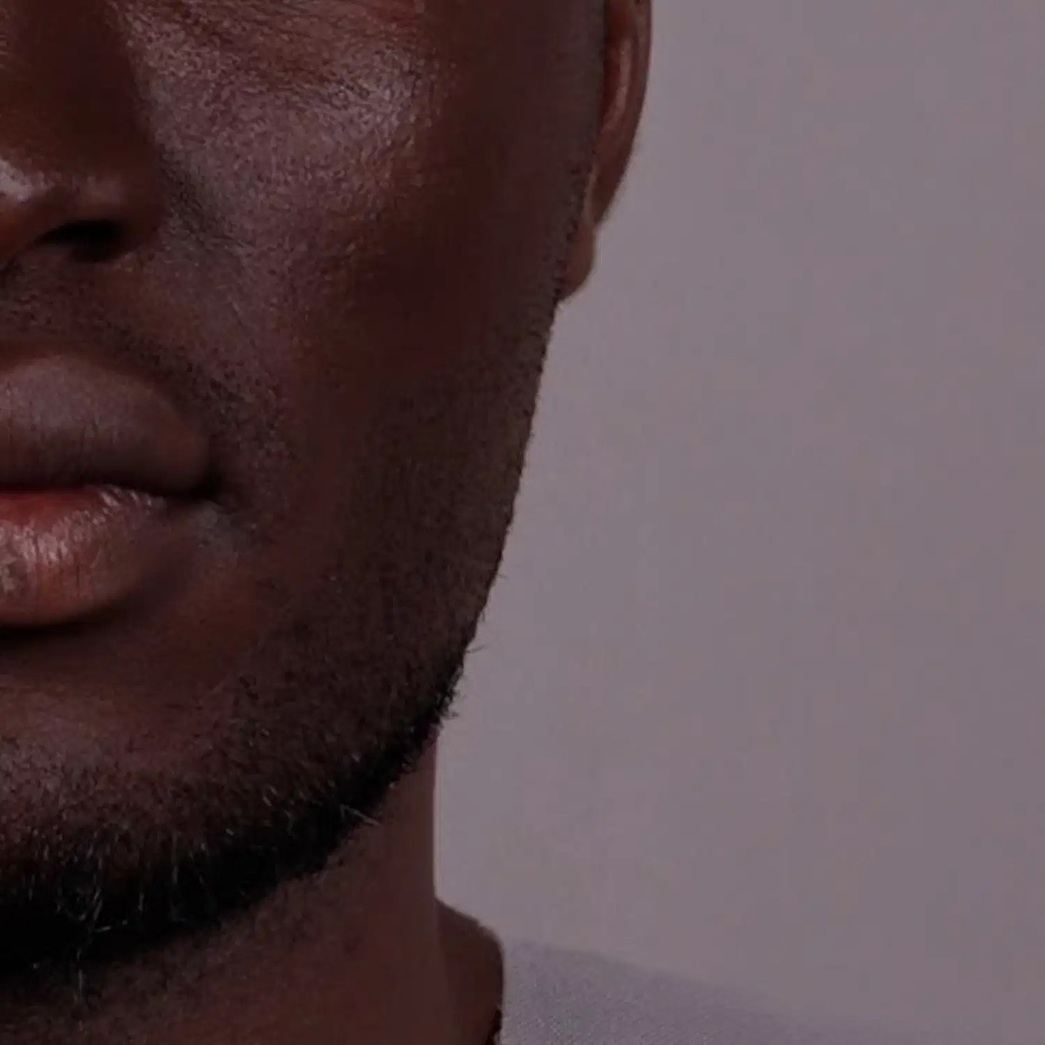 Imagem de close-up do quarto inferior esquerdo de um rosto masculino negro com barba rente e camiseta de gola redonda branca, em um fundo cinza claro.