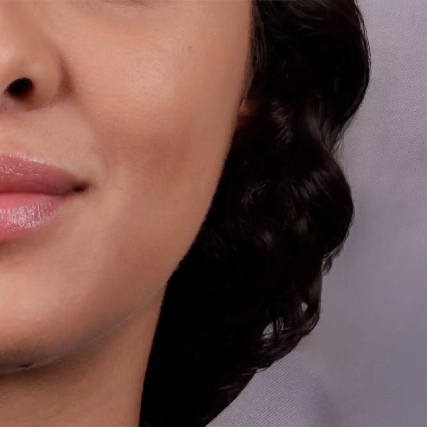 Imagem de close-up do quarto inferior esquerdo de um rosto feminino negro com cabelo longo, cacheado e castanho cobrindo a orelha em fundo cinza claro.