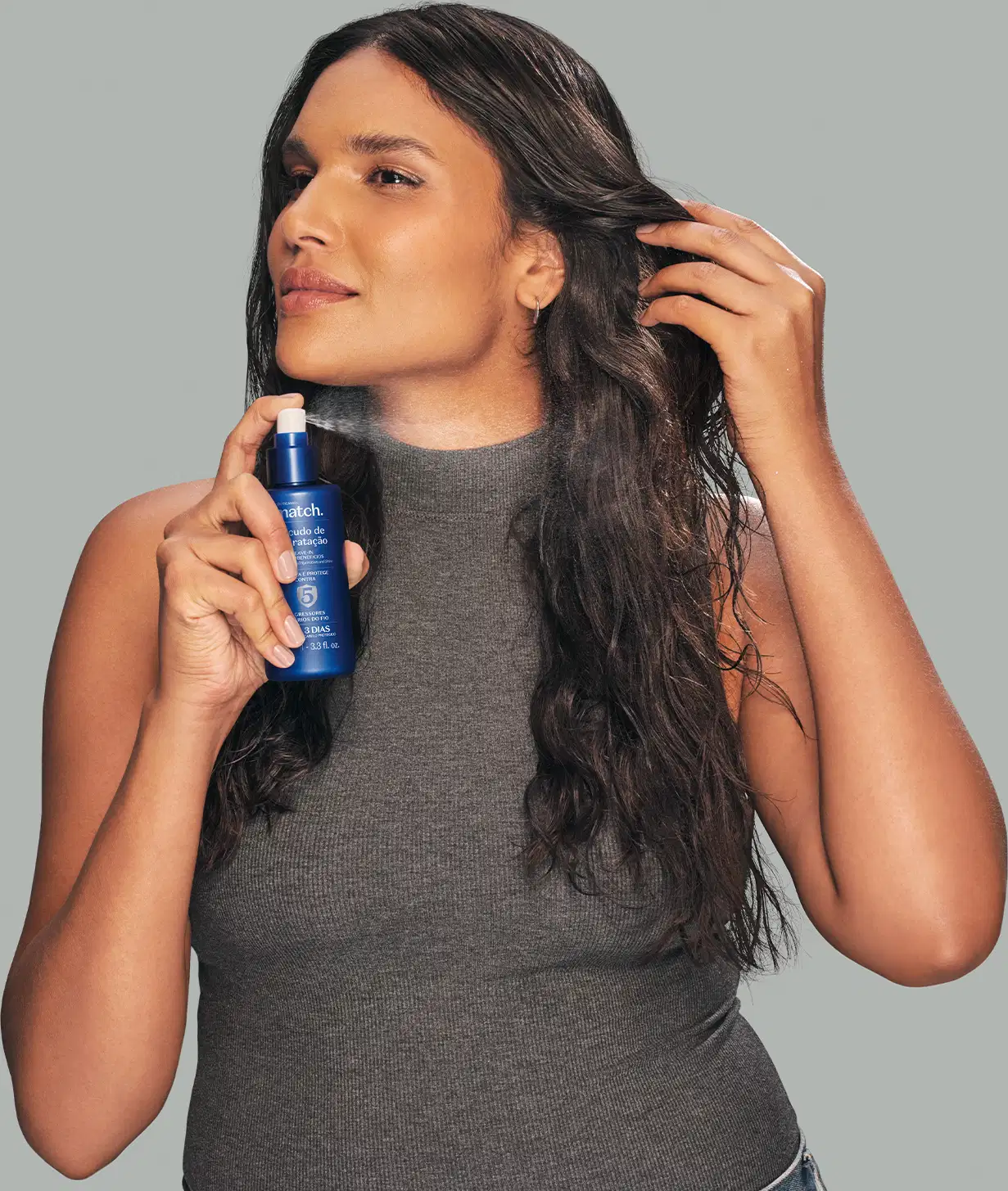 Mulher segurando um item de cabelos azul em frente ao corpo, vestindo uma blusa cinza em um fundo cinza claro. Cabelos longos e ondulados. Ambiente neutro e minimalista que destaca o produto.