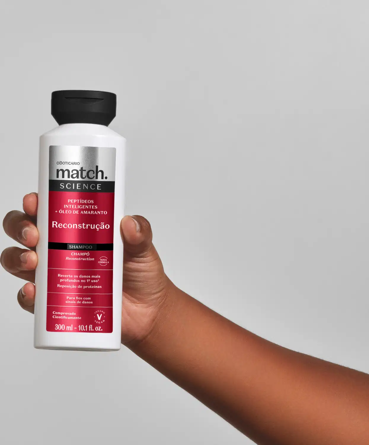 Frasco branco com rótulo vermelho da linha Match Science contendo shampoo para reconstrução capilar, seguro por uma mão feminina em fundo cinza claro.