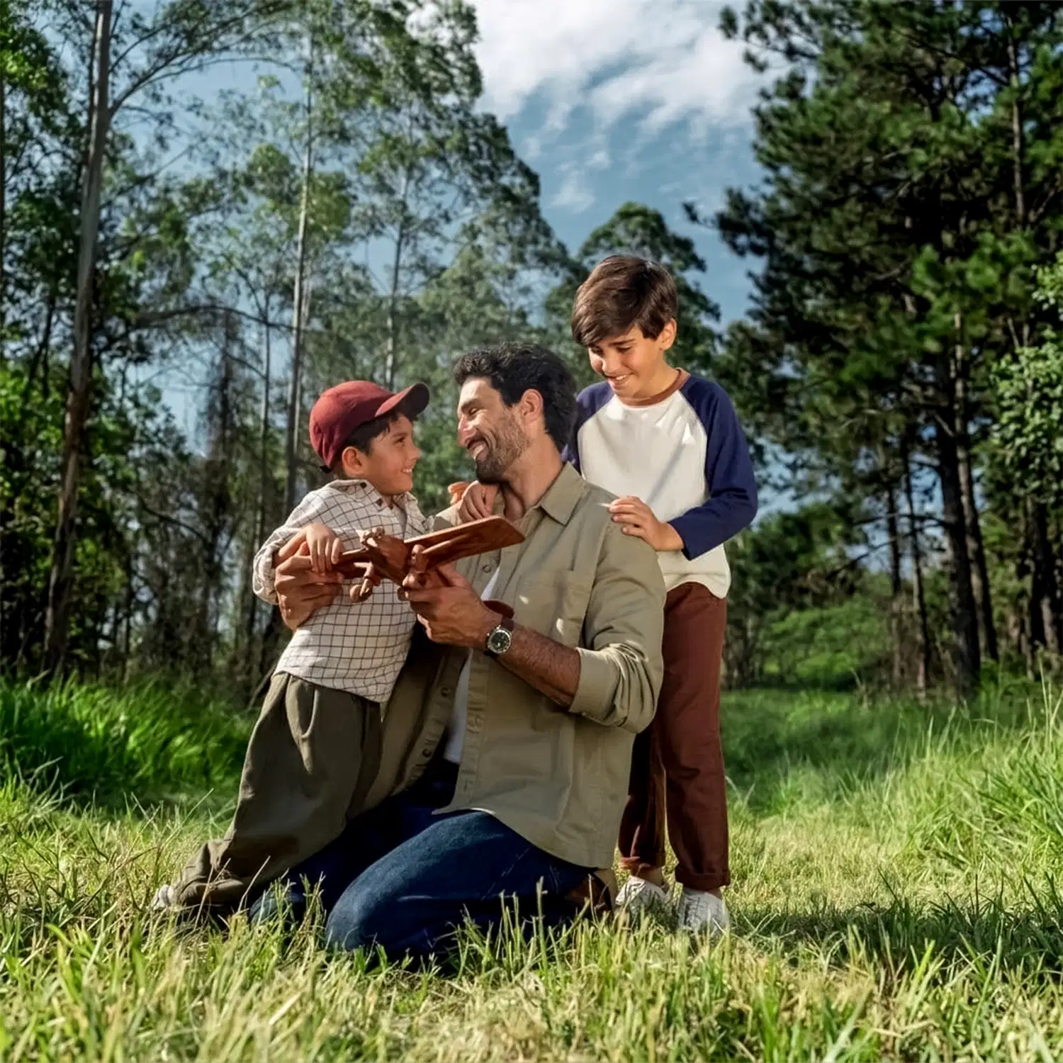 Um homem adulto e dois meninos em um ambiente natural com árvores e vegetação ao redor, interagindo com um livro.