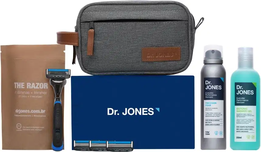 A imagem mostra um kit de produtos Dr. Jones. Há um Cabo de Aparelho de Barbear, 4 lâminas de barbear, uma nécessaire cinza, um frasco de shampoo, um frasco de espuma de barbear cremosa.