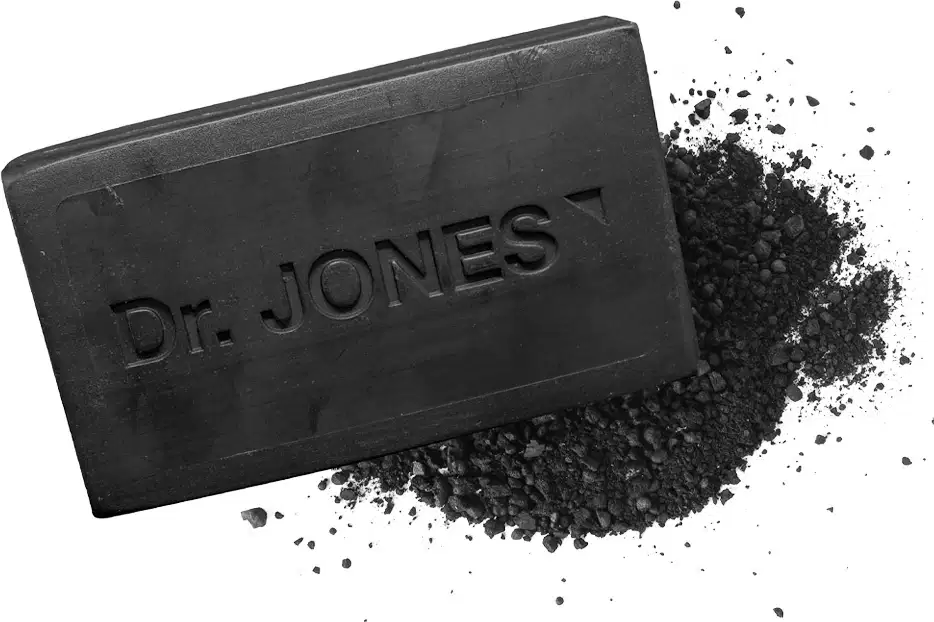 A imagem mostra o sabonete em barra na cor preta com a inscrição "Dr. Jones" em relevo. Ao lado há uma quantidade significativa de carvão espalhado em forma de grânulos.