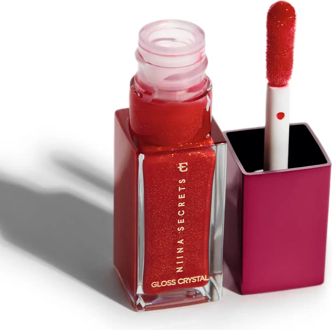 Frasco de vidro retangular transparente contendo um o gloss brilhante vermelho com partículas cintilantes. O frasco está aberto e ao lado está o aplicador labial na mesma cor vermelha cintilante acoplado à tampa compacta de cor rosa escuro.
