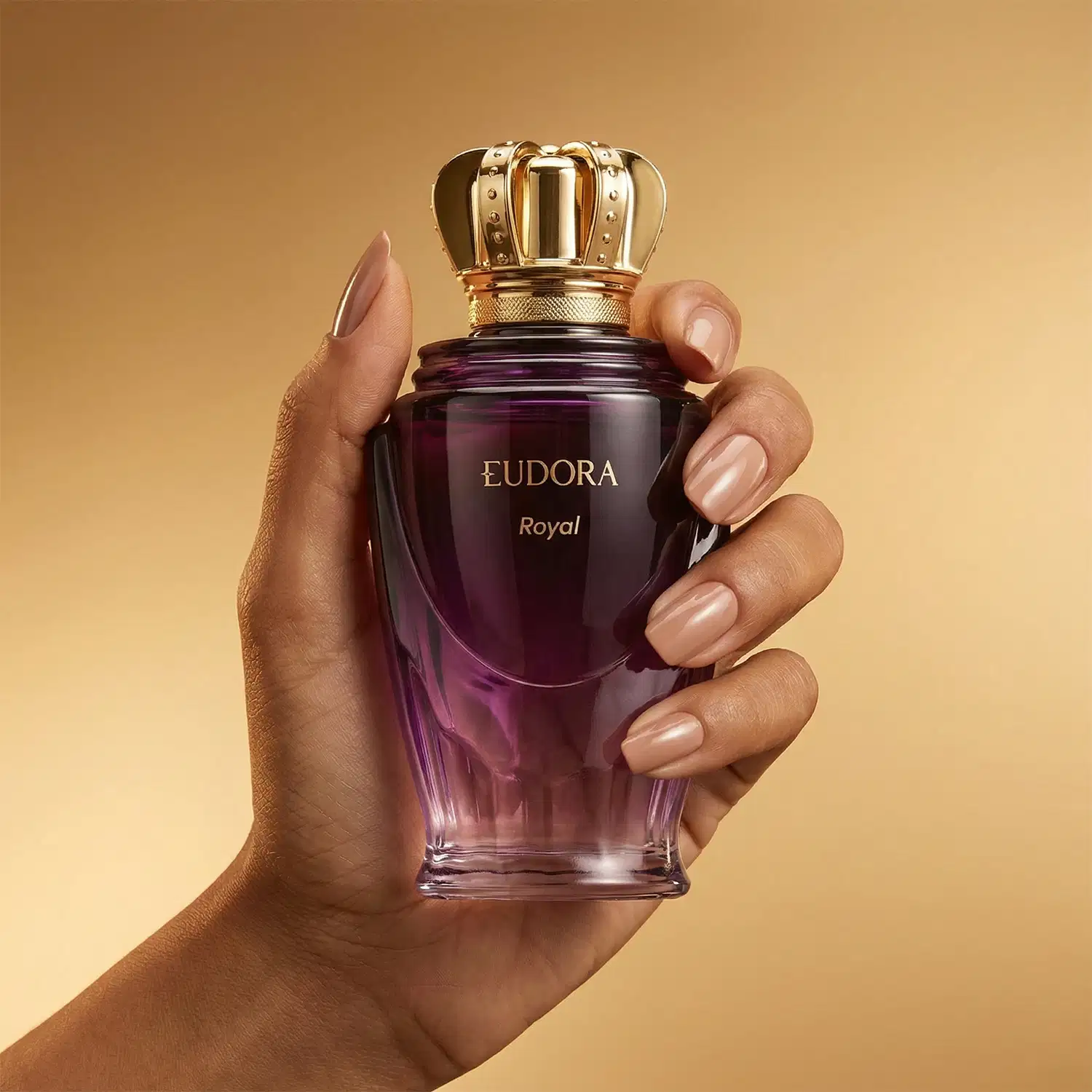 Frasco do Desodorante Colônia Eudora Royal com tampa dourada em formato de coroa, em tonalidades de roxo, segurado por mão feminina com unhas pintadas.