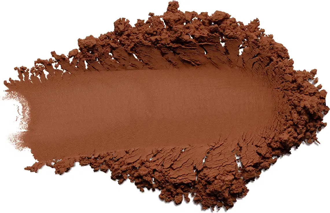 Pó compacto de maquiagem em tom marrom terroso mais escuro cor 6. O produto apresenta uma textura fina e pulverizada, formando uma superfície irregular com pequenos montes de pó solto ao redor de uma área central mais compactada.