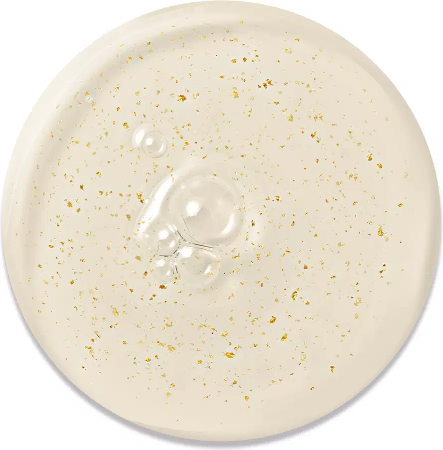 Textura do Primer. Gota bege claro com partículas douradas espalhadas por toda a superfície. Formato circular com diâmetro aproximado de 6 cm. Superfície levemente côncava no centro, onde se concentram algumas gotas de um produto líquido translúcido.