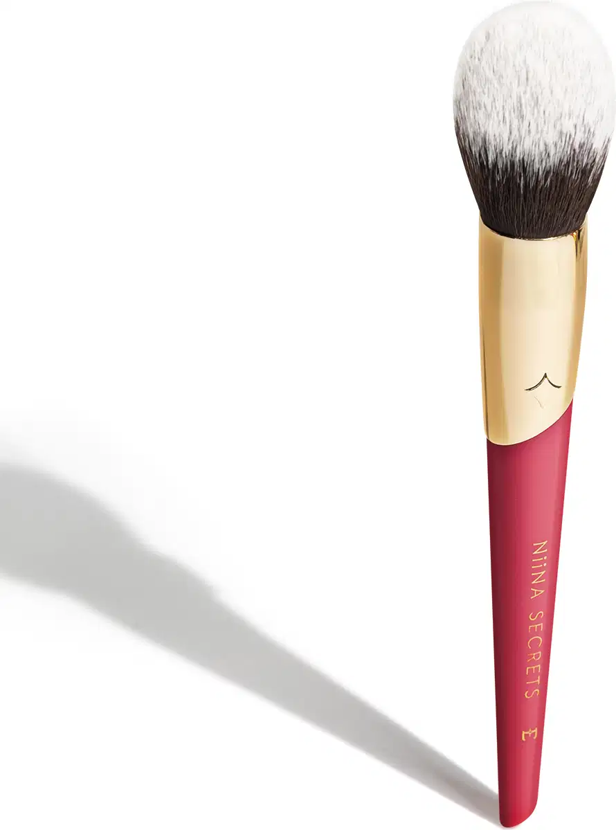 A imagem mostra um pincel de maquiagem, visto de um ângulo um pouco mais elevado, sem ser exatamente de frente. Com cabo vermelho e dourado, e cerdas pretas com ponta branca. Na parte vermelha, logo da marca e linha do produto escrito em dourado.