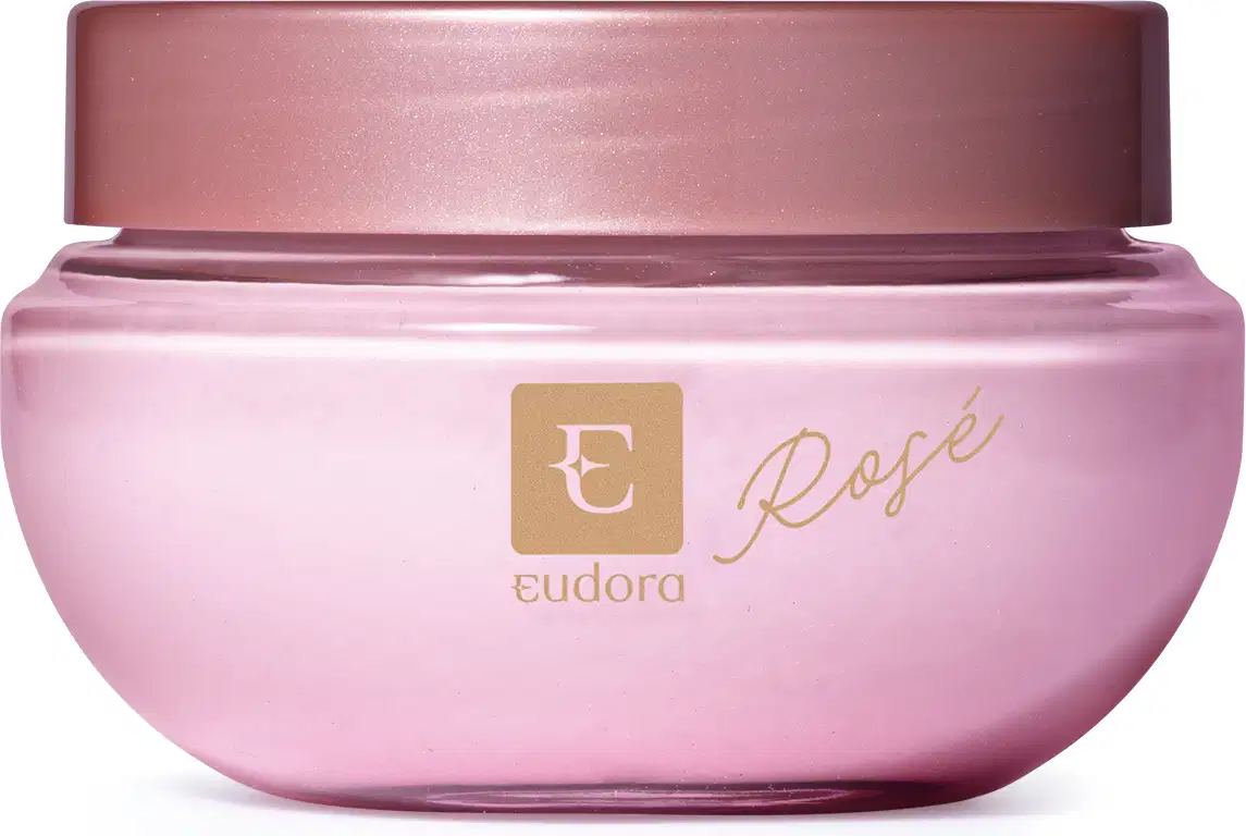 A imagem mostra a embalagem do hidratante Eudora Rosé. É um pote arredondado, rosa claro, com a tampa na cor roxa clarinho. Na parte frontal do pote, está escrito Eudora Rosé em letras douradas e há também o símbolo da marca: um E vazado em fundo dourado.