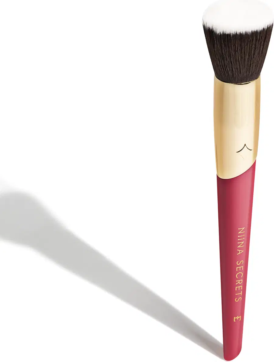 Pincel de maquiagem com cabo rosa e dourado. O cabo é cilíndrico, na cor rosa escura, com o texto "Niina Secrets" impresso. A parte superior é dourada. As cerdas do pincel são densas, corte reto e de cor marrom escura.