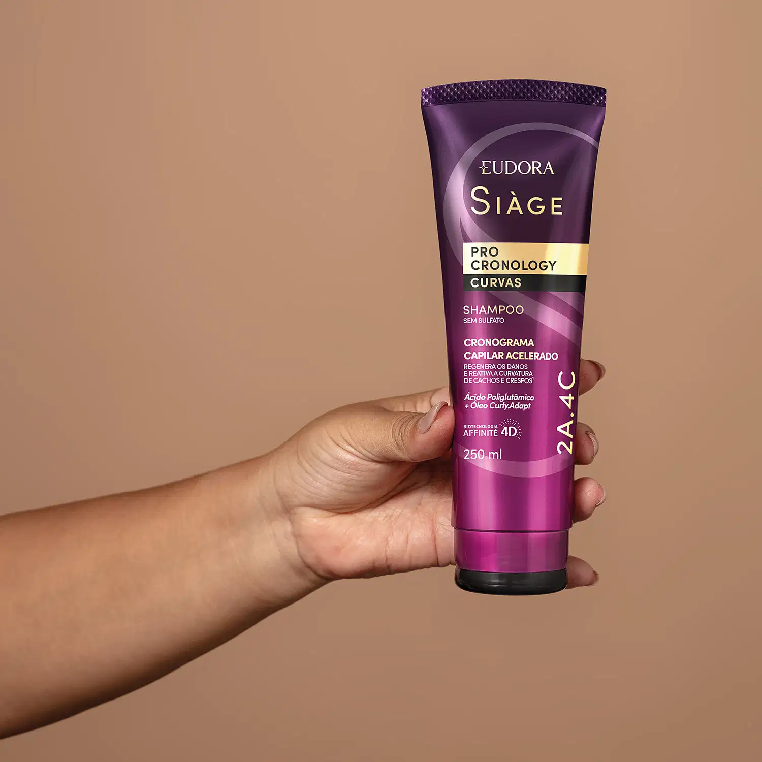 O produto mostrado na imagem está sendo segurado na mão. É um Shampoo da linha Siàge Pro Cronology Curvas, da marca Eudora. O condicionador está em uma embalagem de tubo plástico de cor roxa e rosa, com detalhes texturizados.