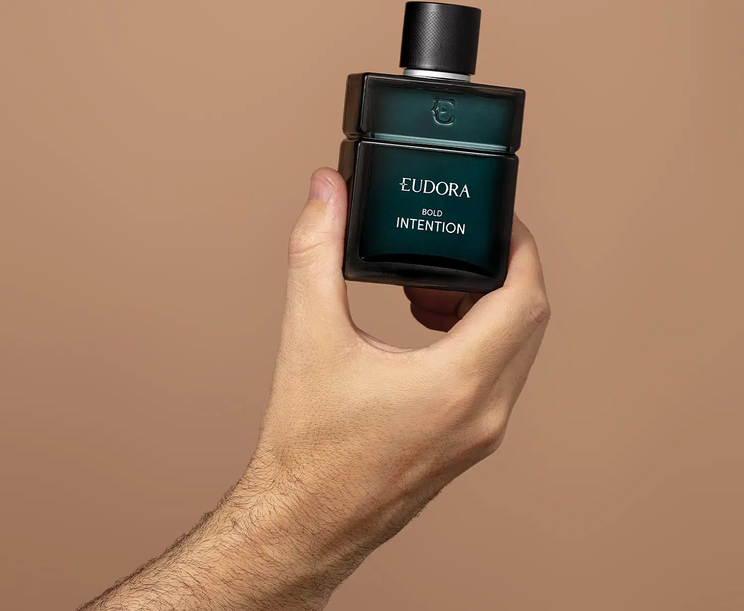 O produto apresentado é um frasco de "Bold Intention". Tem um design moderno e minimalista, com um formato retangular. O produto é segurado por uma mão masculina, sugerindo que se trata de um item masculino da linha "Intention".