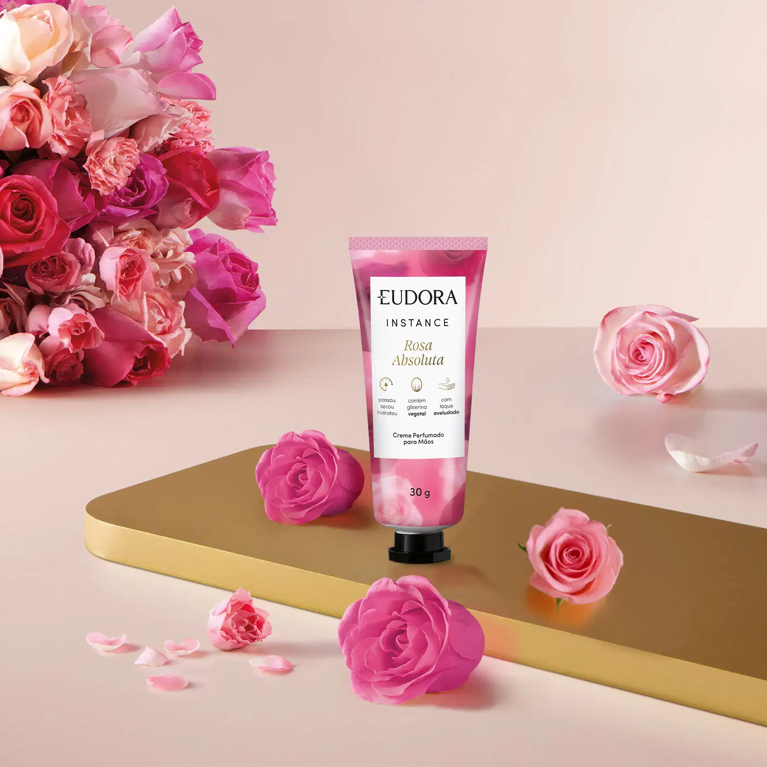 O creme está em uma embalagem de tubo plástico rosa, com o logo da marca e informações sobre os ingredientes. O produto está posicionado em uma superfície dourada, com algumas rosas frescas e pétalas espalhadas ao redor, criando uma composição floral.
