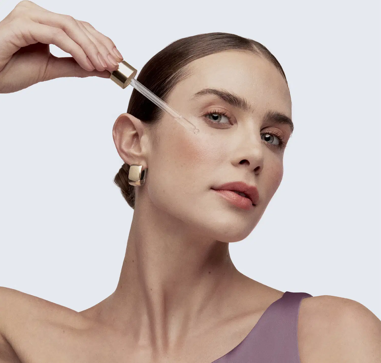 Uma mulher aplica um produto de skincare em seu rosto, com uma conta gotas transparente, com tampa dourada em primeiro plano.