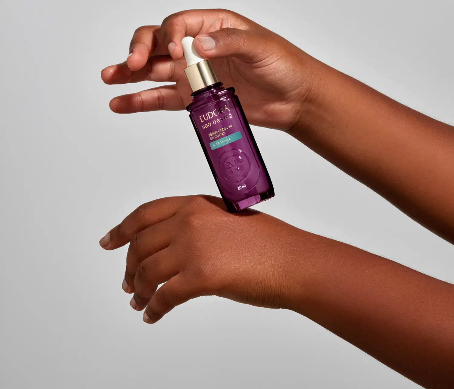 Frasco roxo com rótulo dourado da marca Eudora Neo Dermo, com uma tampa em conta gotas. O produto está sendo segurado por mãos femininas.